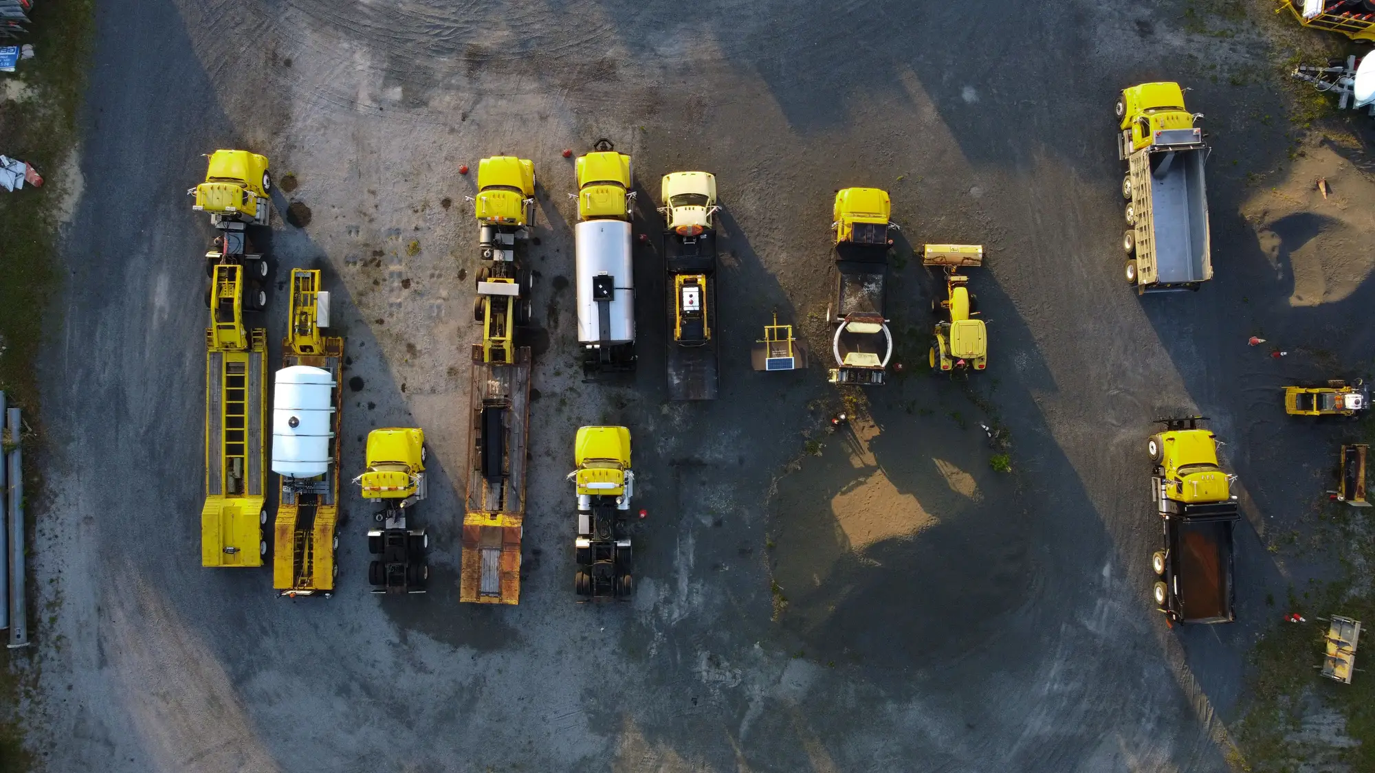A gravel equipment yard seen from straight above: yellow trucks, trailers and a tanker parked in rows, with a few smaller machines off to the side